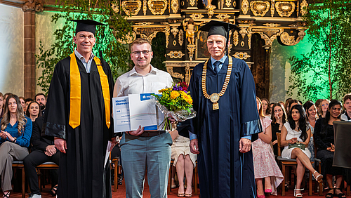 Julius Seifert: Absolvent mit Preis, Laudator Professor Jan Thomanek (l.) und Rektor Professor Volker Tolkmitt. Drie maännliche Personen stehend im Altarrarum einer Kirche. Die jünger Person in der Mitte hält einen Blumenstrauß und eine Urkunde in den Händen. Die Personen links und rechts tragen akademsiche Talare und Hüte, die Person rechts zusätzllich eine goldene Amtskette.